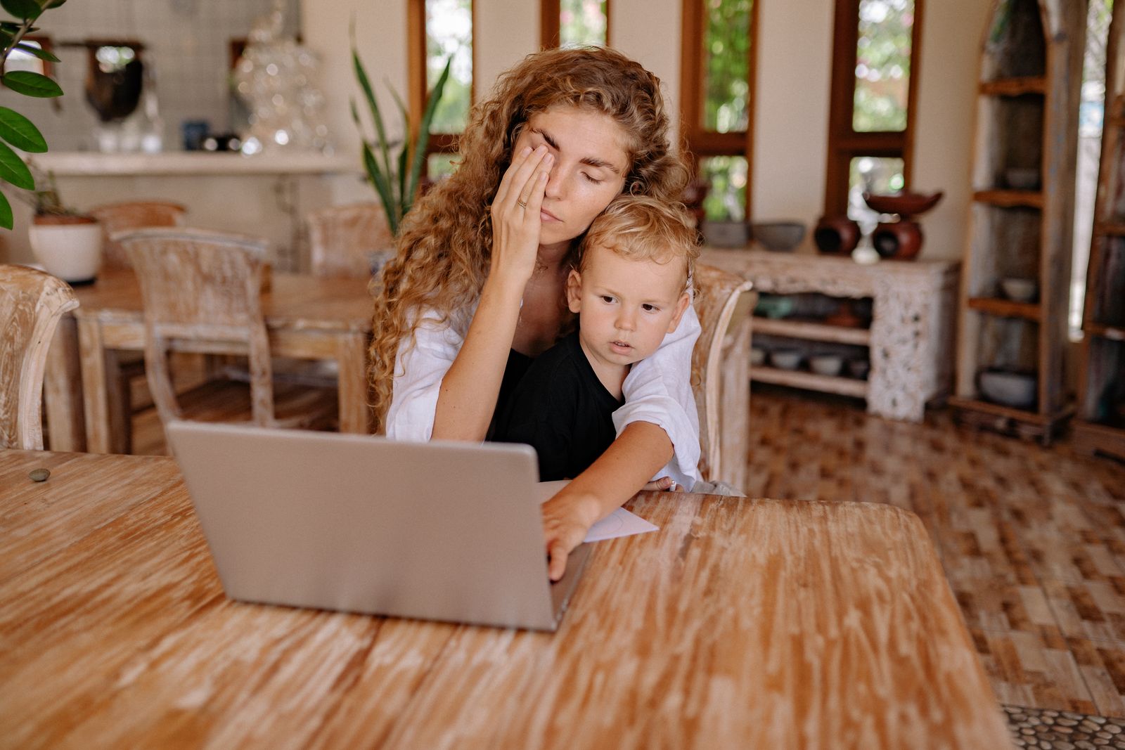 Une mère qui travaille sur son ordinateur pendant que son fils s'appuie contre elle, fatiguée mais présente